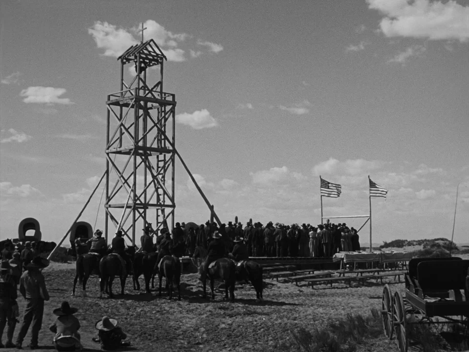 My Darling Clementine (1946) movie still: wide — A wooden bell tower stands in the center of a desert gathering site. A large crowd of people in…
