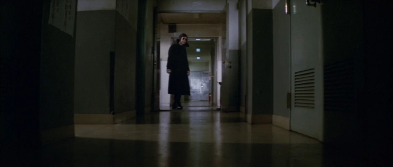 Godzilla Vs Mechagodzilla (1974) movie still: wide — A dimly lit hospital hallway with dark walls leads to a brighter open doorway. A man in a long…