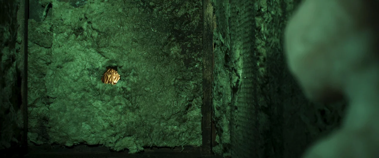 Stopmotion (2024) movie still: closeup — A dark corner is filled with thick, matted green fiberglass building insulation. A small hole in the…