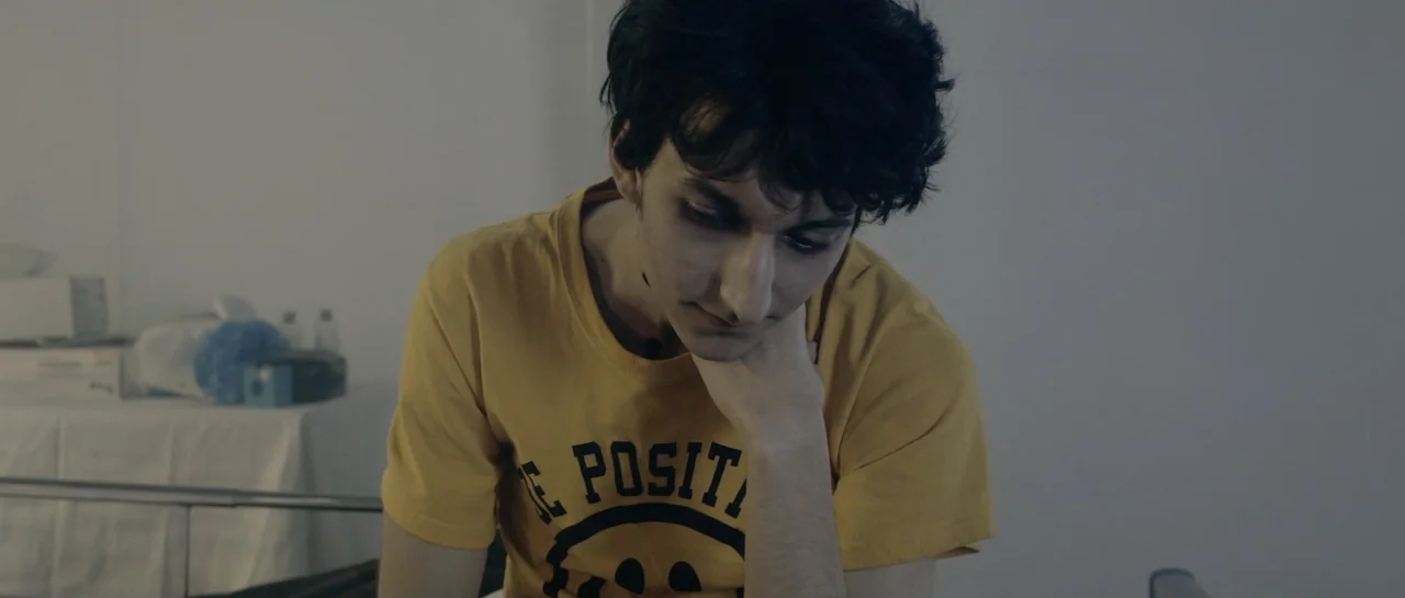 NY84 (2016) movie still: medium — A spare, neutral-colored indoor space features a young man against a plain white wall. The subject sits in the…
