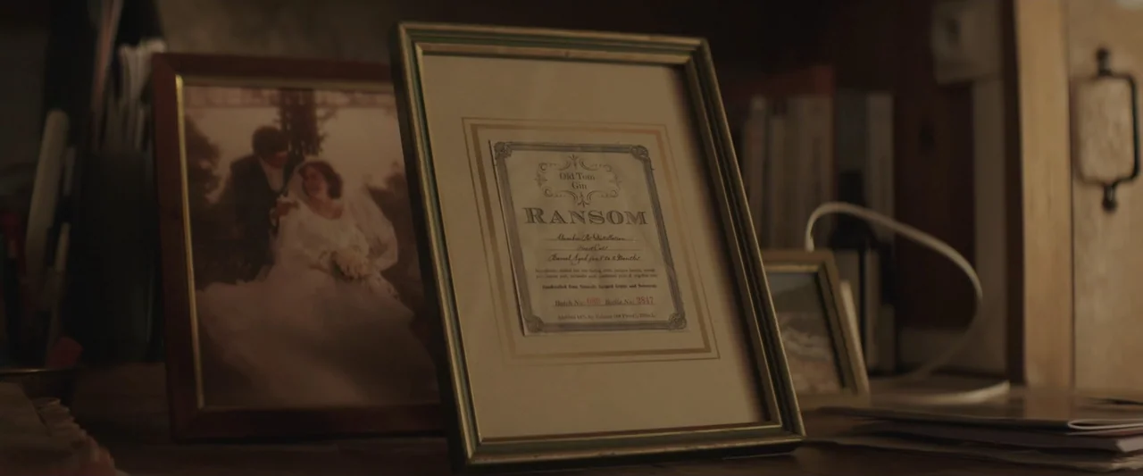 The Only One (2021) movie still: closeup — A wooden desktop surface holds a framed gin label and a framed wedding photo. Items are arranged casually…
