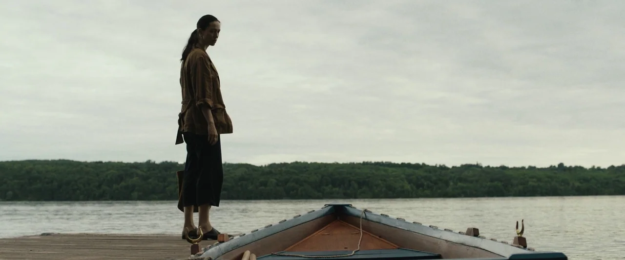 The Night House (2020) movie still: fullbody — A woman stands on a wooden dock next to a small wooden boat. The background shows a wide river with a…