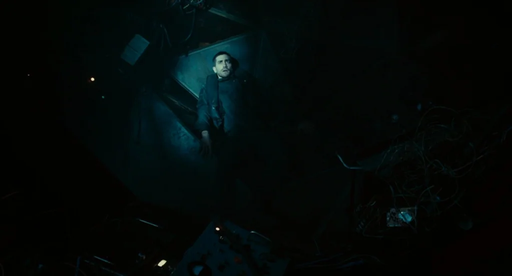 Source Code (2011) movie still: fullbody — A man lies inside a dimly lit, narrow metallic container surrounded by wires and debris. Cables hang from…