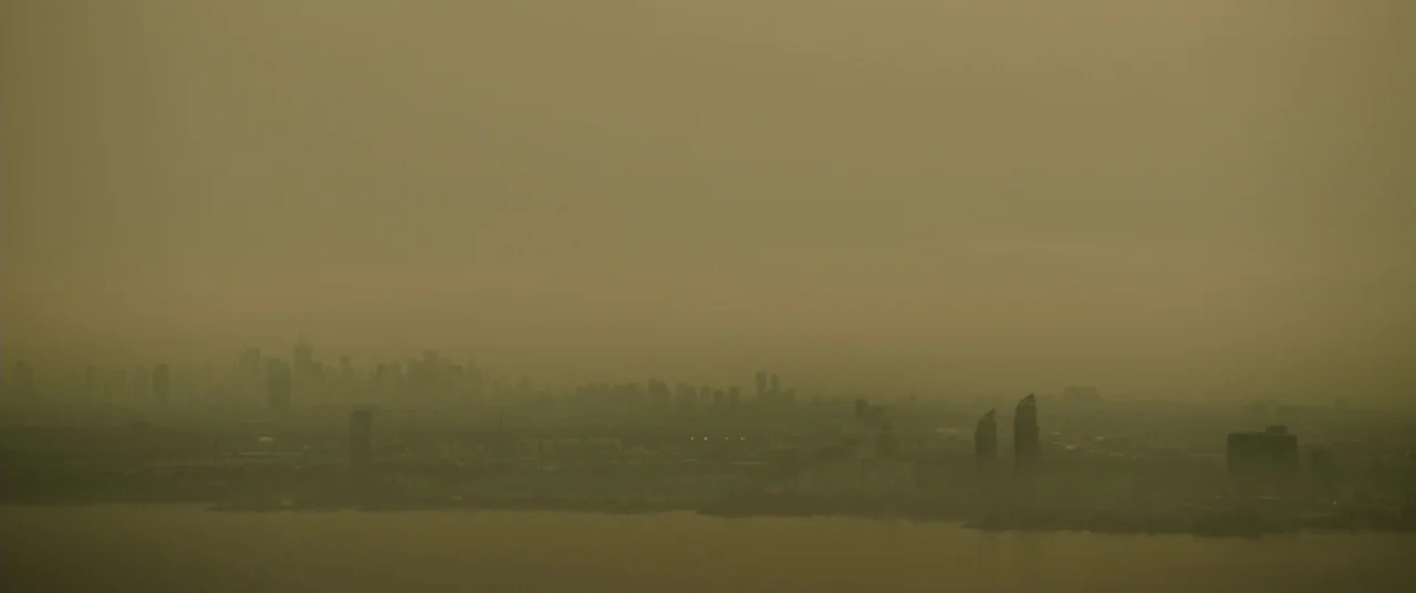 Enemy (2013) movie still: establishing — A distant urban skyline emerges faintly through dense smog above a body of water. Tall building silhouettes…