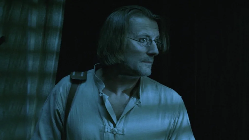 The Ugly Swans (2006) movie still: medium — A man stands in a dark room illuminated only by a side light source. He wears thin glasses and gazes…