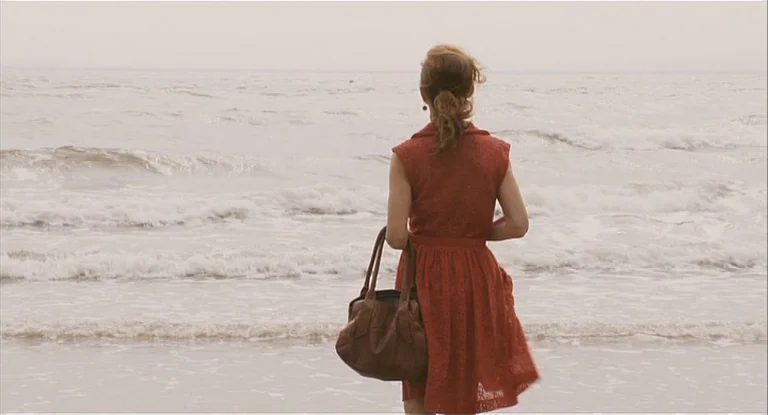 In Another Country (2012) movie still: fullbody — A wide sandy beach under a flat, overcast sky meeting the surf. A woman in a red dress stands…