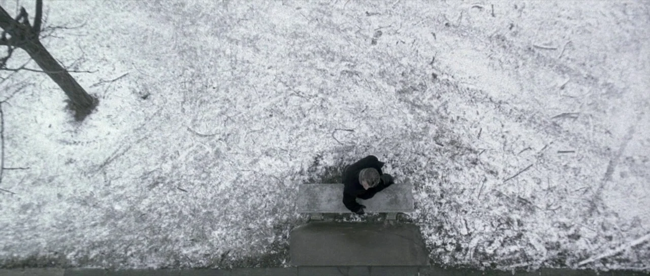 The Mothman Prophecies (2002) movie still: wide — A top-down view of a snow-covered park ground with a single stone bench. An elderly man in a black…