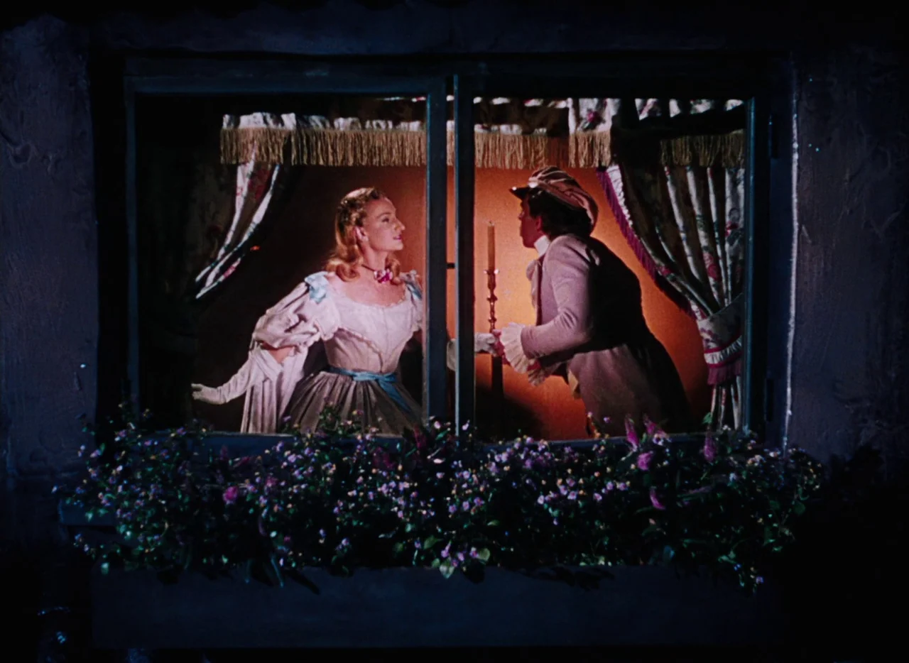 The Tales of Hoffmann (1951) movie still: medium — A dark exterior view of a window illuminated from within by a single candle. A man and a woman…