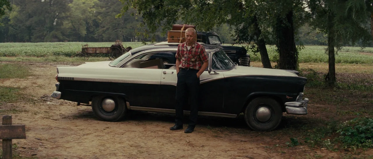 Loving (2016) movie still: fullbody — A rural landscape features a dirt path adjacent to a large green field. A man with a suitcase atop his car…