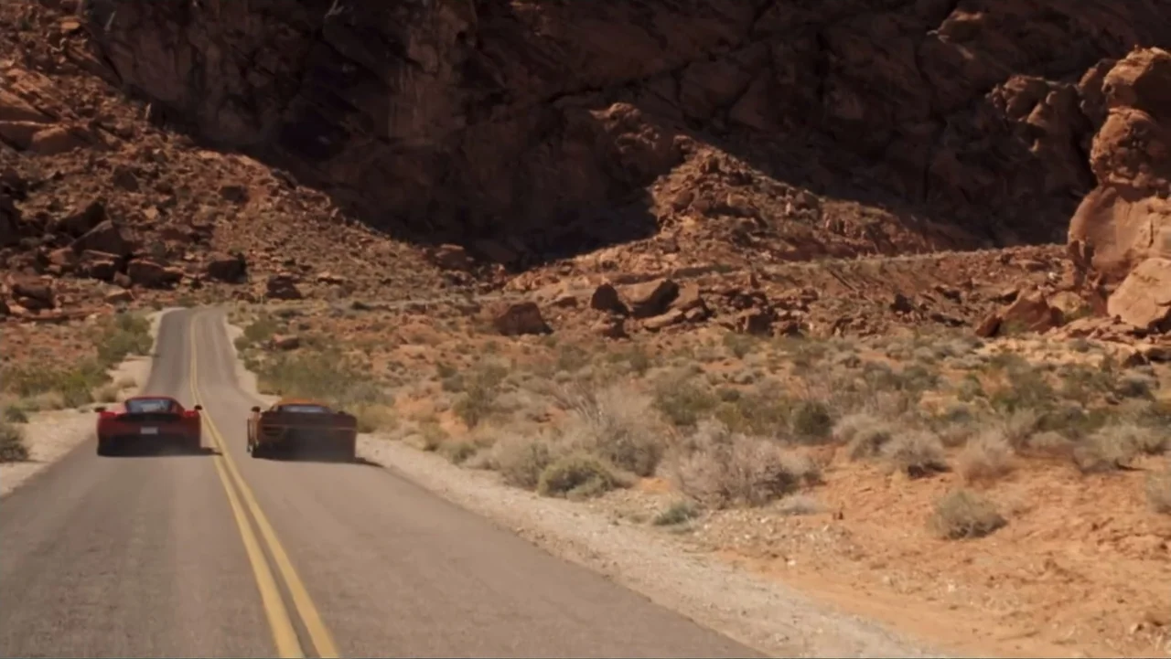 Redline (2007) movie still: wide — A remote two-lane road winds through a rocky desert landscape with sparse scrub brush. Two sports cars travel…