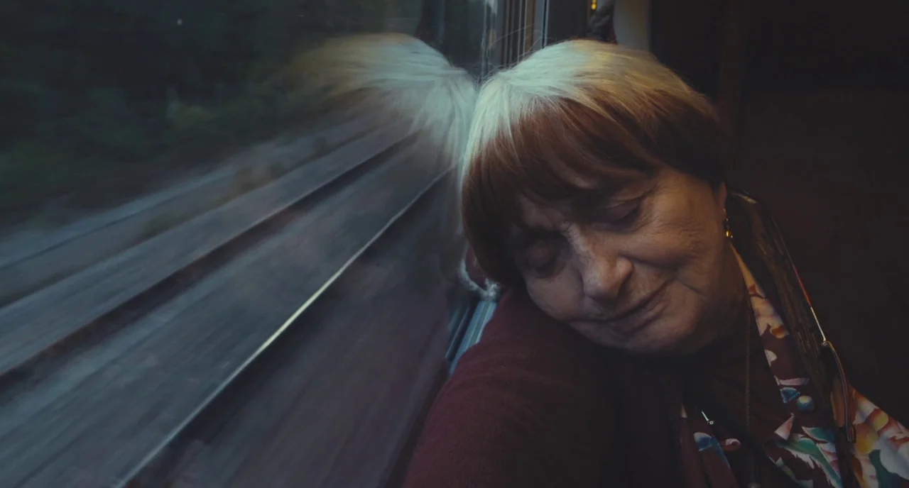Faces Places (2017) movie still: closeup — An elderly woman sits inside a moving train with her head against the glass. Outside the window, the…