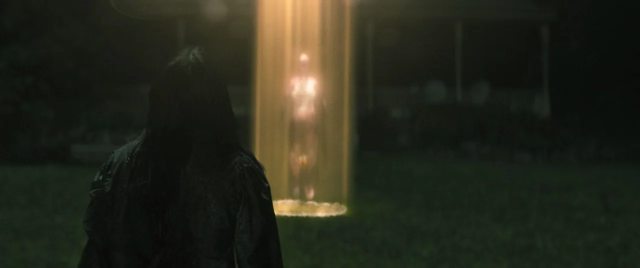 No One Will Save You (2023) movie still: medium — A dark, grassy yard at night is illuminated by a singular, vertical column of light. A person…