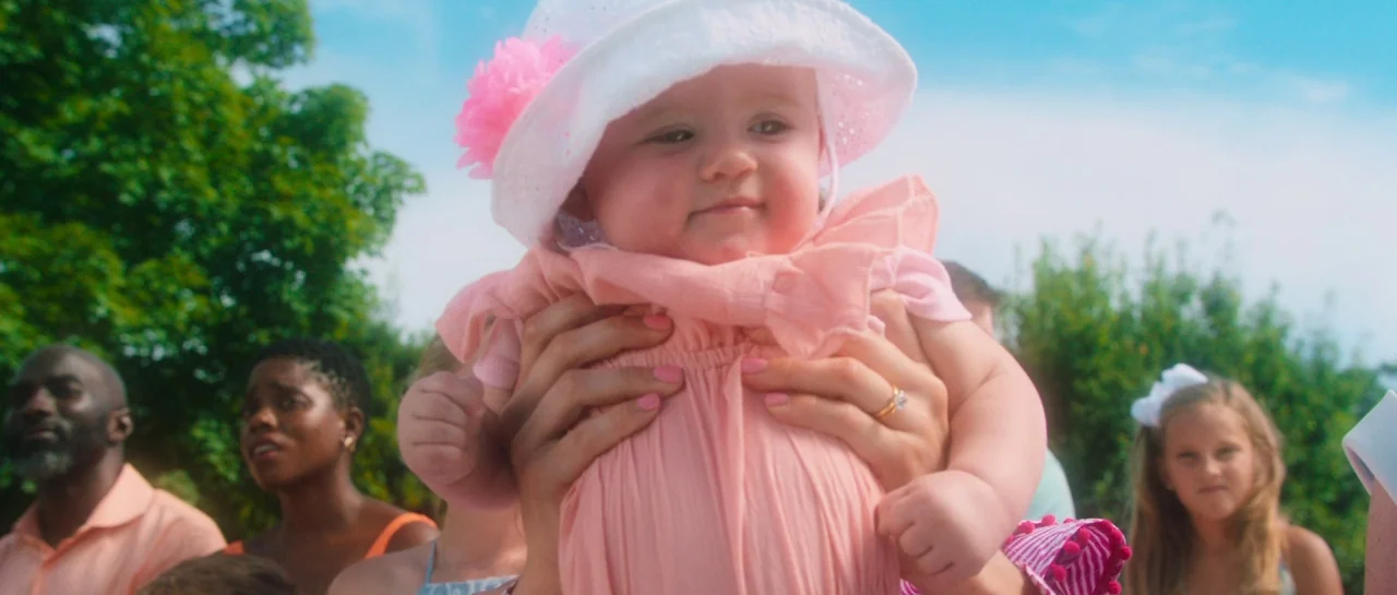 Greener Grass (2019) movie still: closeup — An outdoor park setting with lush green trees and a clear blue sky. An infant in a pink dress and sun…
