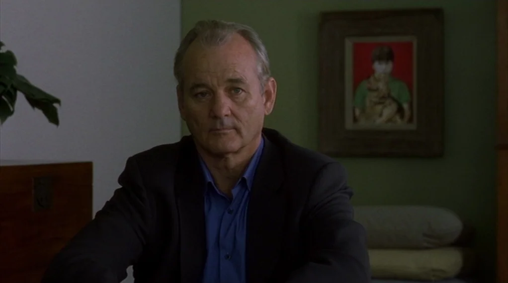 Broken Flowers movie still: medium — A man sits in front of a green wall with a framed portrait behind him. He stares forward while positioned in a…
