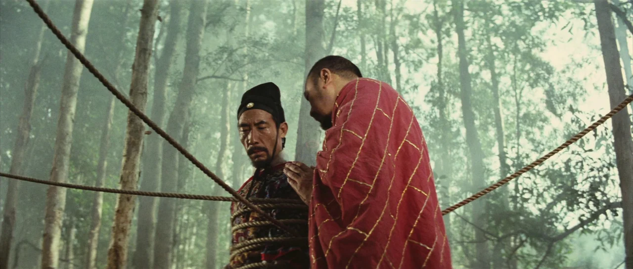 A Touch of Zen (1971) movie still: medium — A man is tied to trees in a dense, hazy temperate forest setting. A Buddhist monk in red robes stands…