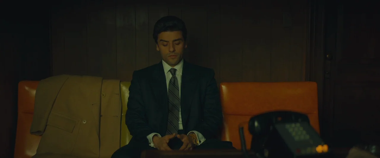 A Most Violent Year (2014) movie still: medium — A dimly lit room with wood-paneled walls features orange chairs and a telephone. A man in a suit…