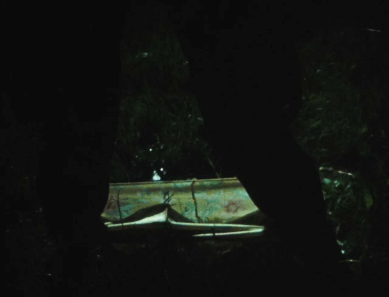 Nekromantik (1988) movie still: closeup — An unidentified metallic object rests on damp grass at night. Large dark silhouettes obscure the…