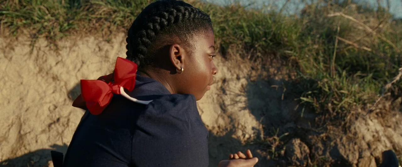 All Dirt Roads Taste of Salt (2023) movie still: medium — A girl with braided hair and a red bow sits on a grassy hillside. The landscape features a…