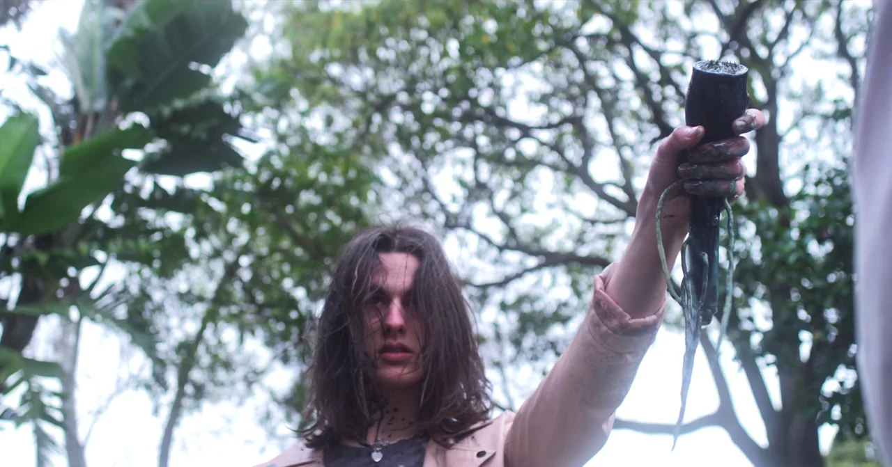 T Blockers (2023) movie still: medium — A person stands among dense trees with blurred branches in the background. The individual holds up a dark…