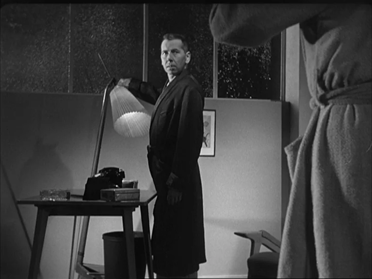 The Sleeping Tiger (1954) movie still: medium — An apartment interior features a desk with a lamp and a window behind. A man stands at the desk and…