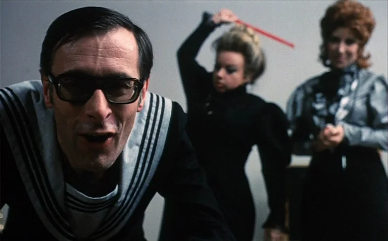 Business is Business (1971) movie still: medium — Three individuals stand before a plain, light grey wall in an indoor space. A man in a sailor suit…
