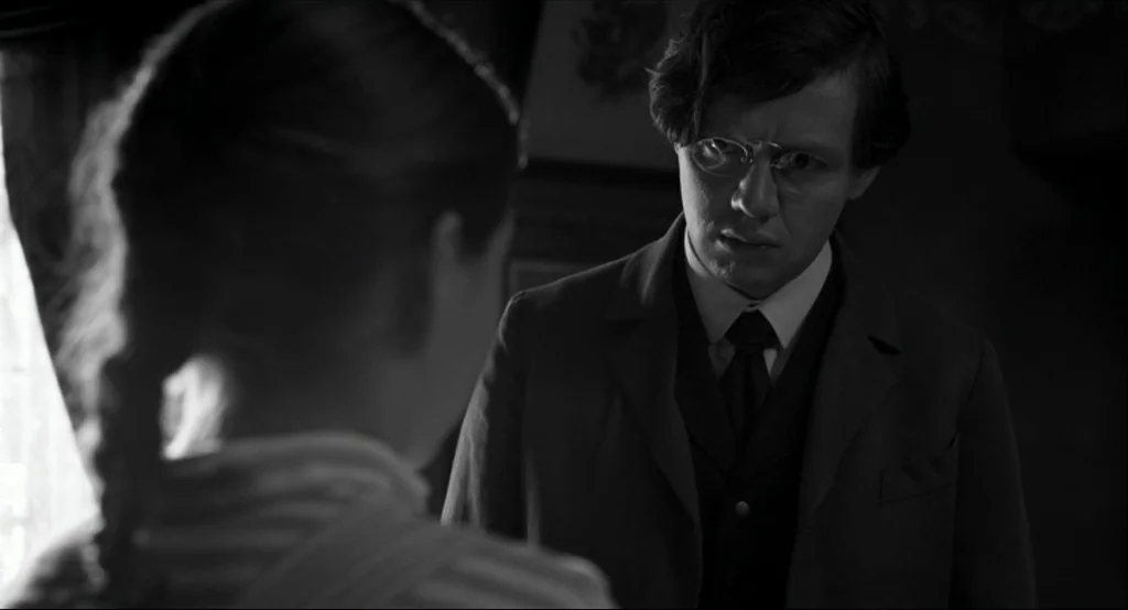 The White Ribbon (2009) movie still: medium — A dimly lit room with traditional furniture visible in the soft-focused background. A man in formal…