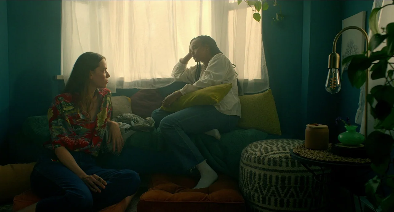 Trim Season (2023) movie still: medium — A cozy corner space features dark teal walls, a draped window, and varied floor cushions. Two young women…