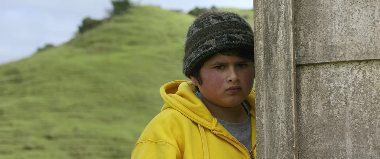 Hunt for the Wilderpeople (2016) movie still: medium — A young boy wearing a yellow hoodie peeks around the corner of a wall. Rolling green hills…