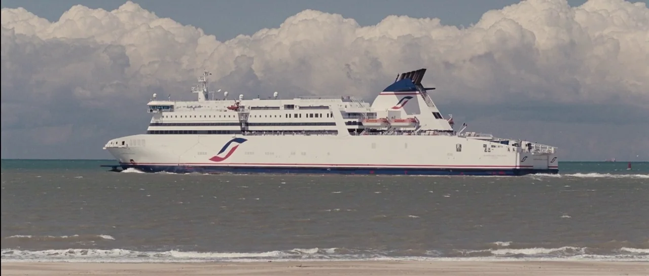 Our Day Will Come (2010) movie still: wide — A large commercial ferry cruises across calm coastal waters under a bright cloudy sky. The vessel is…