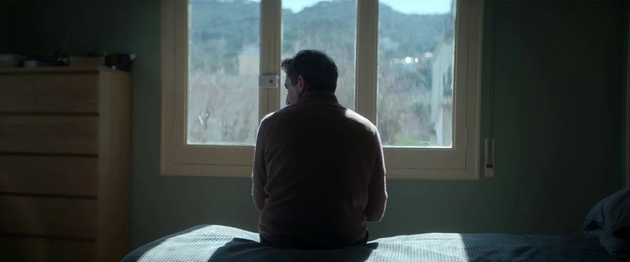 Crazy About Her (2021) movie still: medium — A man sits on a rumpled bed in a dimly lit room with muted green walls. He is turned away from the…