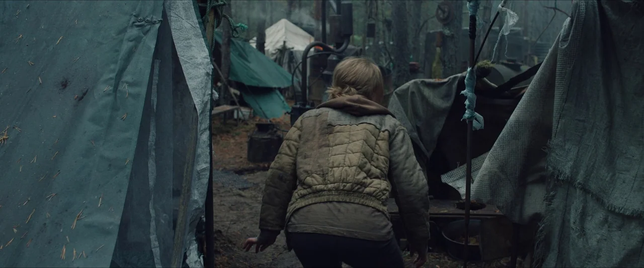 Azrael (2024) movie still: medium — A makeshift encampment is nestled within a dense, misty woodland setting. A young woman walks through the camp,…