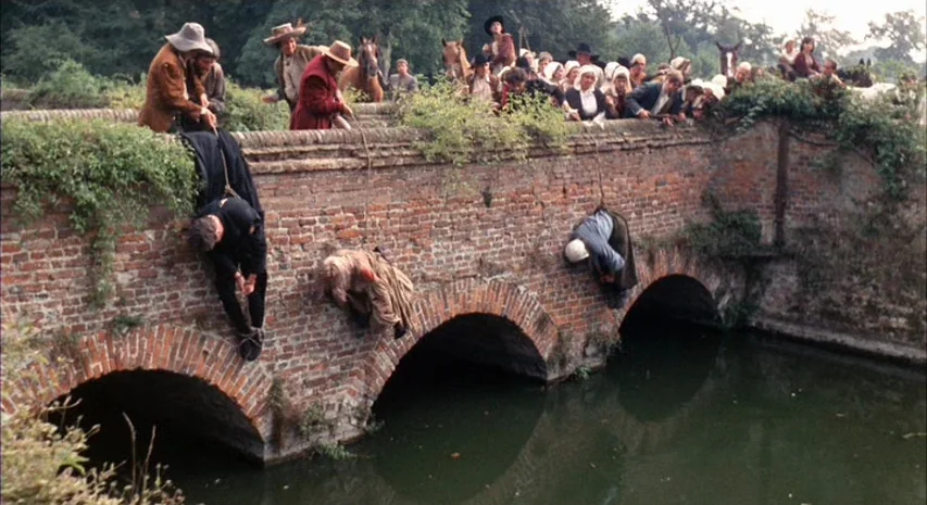 Witchfinder General (1968) movie still: wide — An aged brick bridge spanning a murky stream is crowded with onlookers on top. Three individuals are…