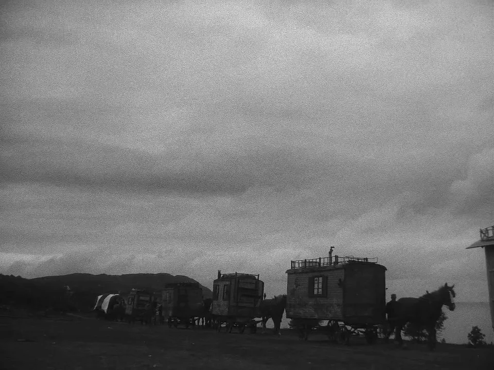 Sawdust and Tinsel (1953) movie still: wide — A series of wooden wagons pulled by horses travel along a dirt path. A vast overcast sky covers the…