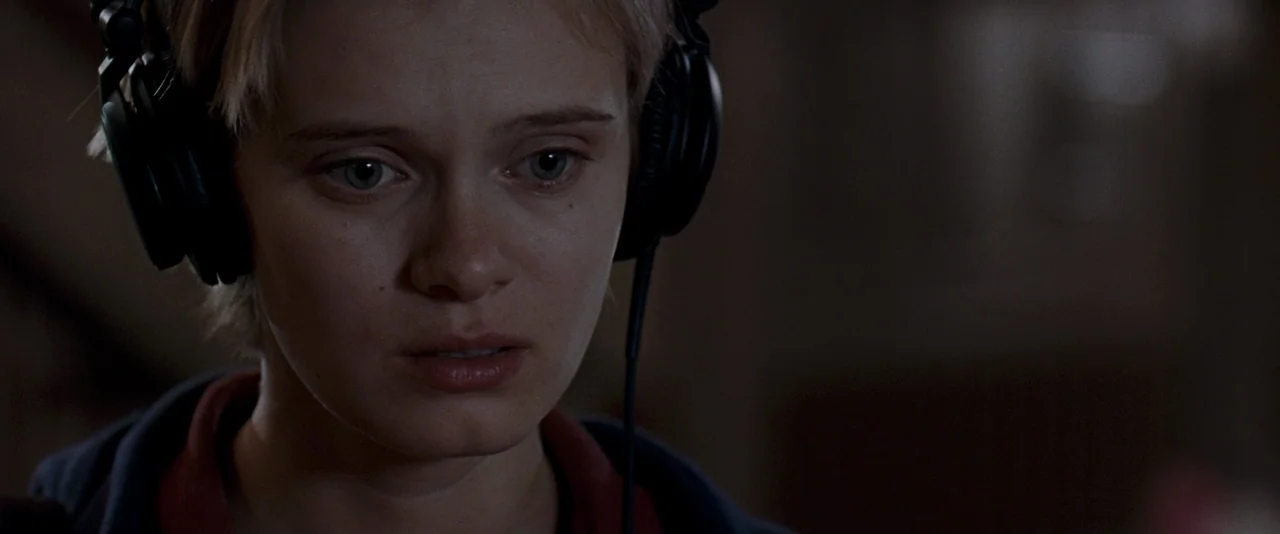 The Innkeepers (2011) movie still: closeup — A dimly lit indoor space with a blurred, dark background. A young woman wears black headphones while…