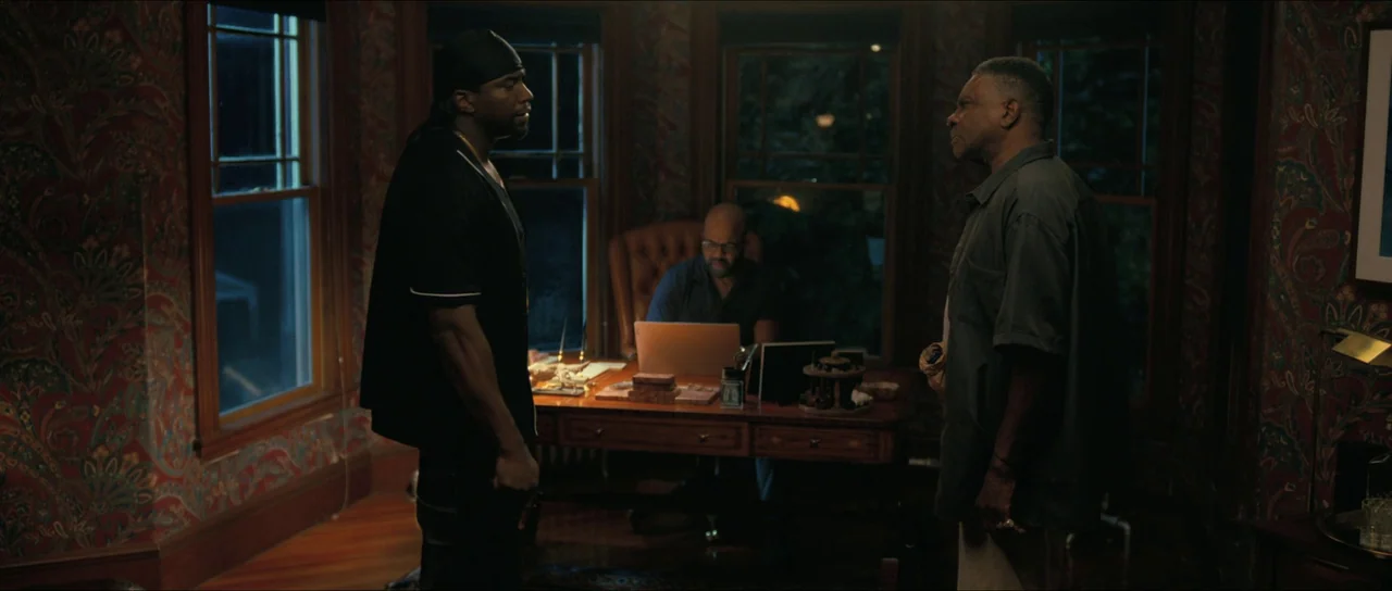 American Fiction (2023) movie still: medium — A dimly lit room features patterned red wallpaper and wooden window frames at night. Three men…