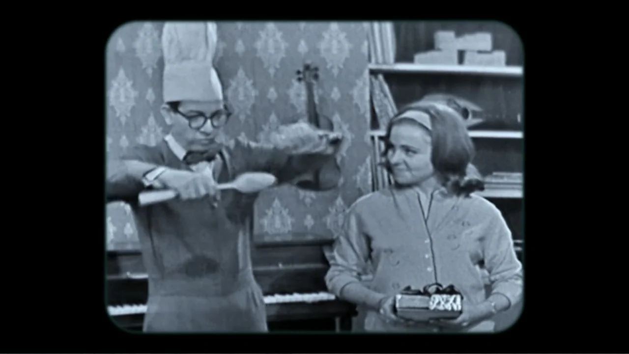 Mr. Dressup: The Magic of Make Believe (2023) movie still: medium — A 1950s living room features patterned wallpaper, a piano, and shelves filled…