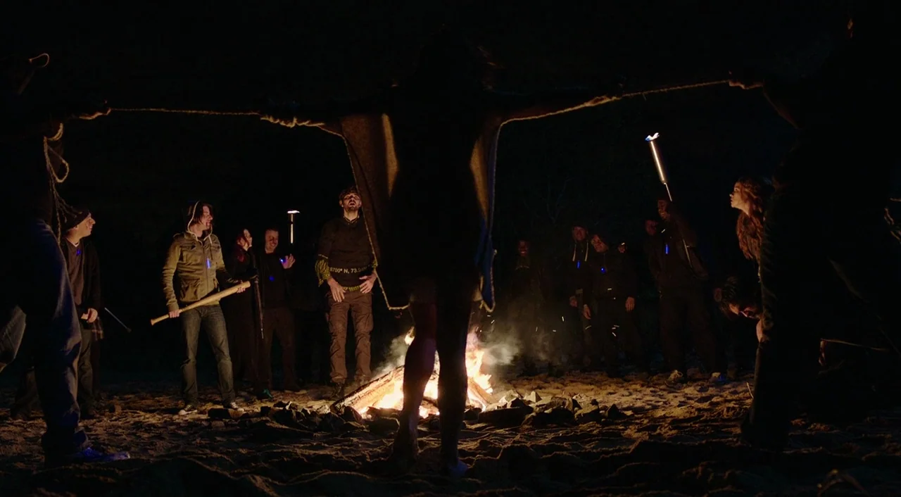Urge (2016) movie still: fullbody — A group of people surround a fire on a dark sandy shore at night. A central figure stands as a silhouette while…
