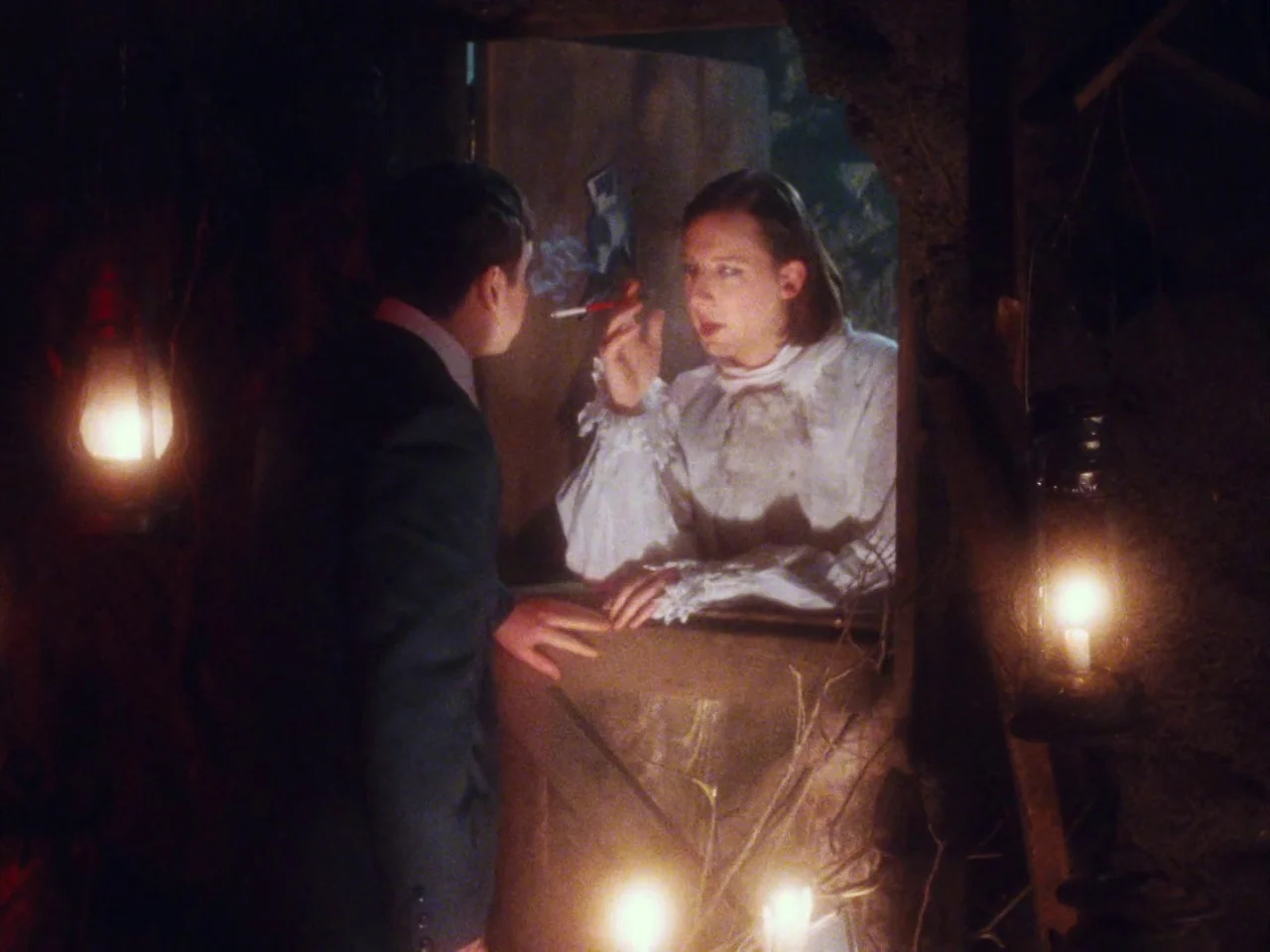 The Twentieth Century (2019) movie still: medium — A dimly lit room with rough walls and multiple hanging lanterns. Two individuals engage in…