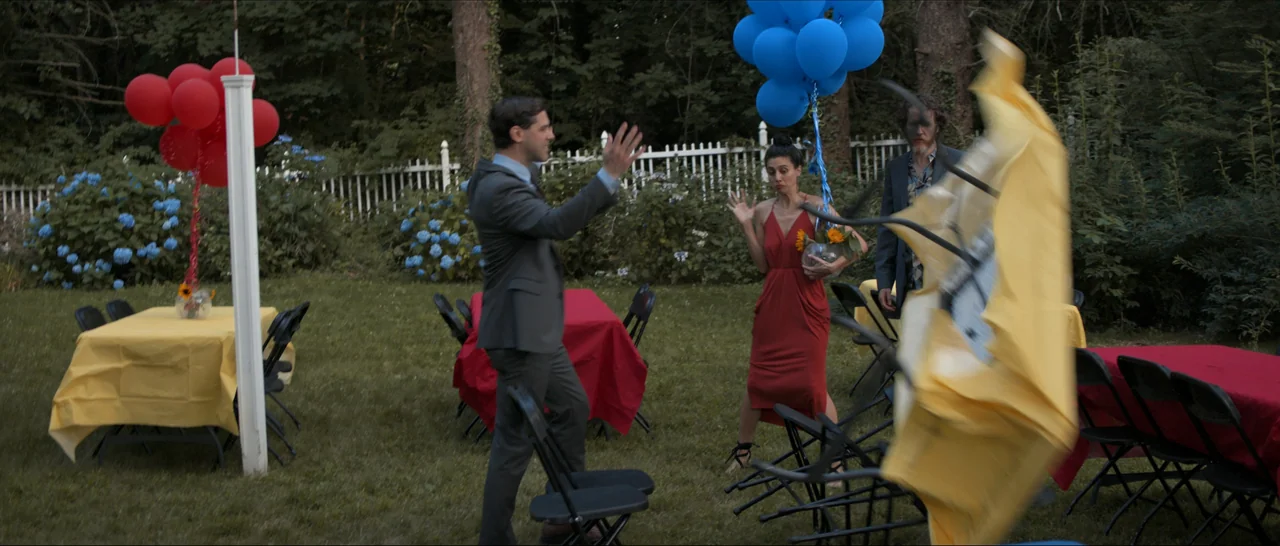 Songs For A Sloth (2021) movie still: wide — A backyard party setting features folding tables with yellow and red tablecloths on grass. A man…