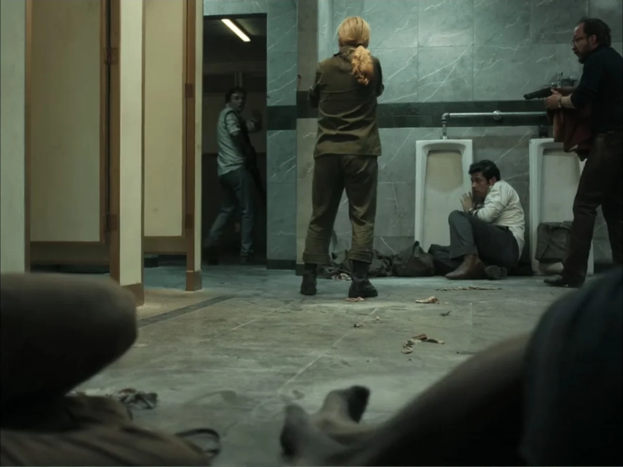 November (2025) movie still: fullbody — A public restroom features grey tiled walls and several ceramic urinals mounted on walls. A woman in…
