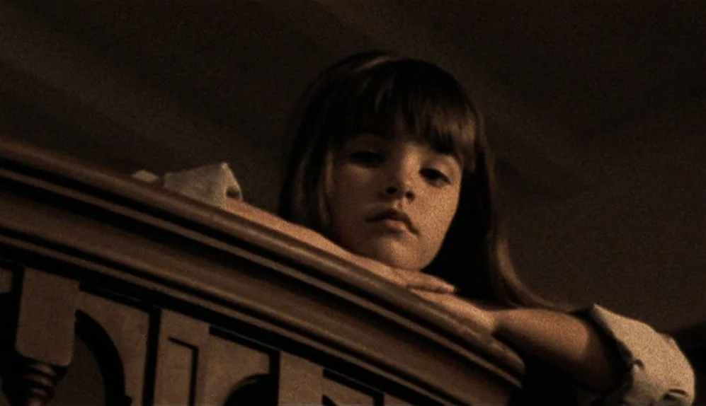 The Limey (1999) movie still: closeup — A dark, interior space features a detailed wooden bannister in the foreground. A young girl rests her chin…