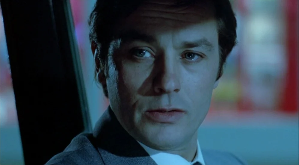 Un Flic (1972) movie still: closeup — The interior of a moving vehicle at night with blue ambient light. A man in a suit looks ahead with a focused…