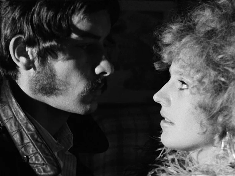 Gods Of The Plague (1970) movie still: closeup — Two individuals stand in a dimly lit, high-contrast black and white space. The couple faces each…