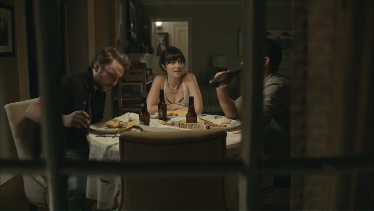 The Hollars (2016) movie still: medium — A residential dining room at night viewed through a glass panel door frame. Two men and a woman sit at a…