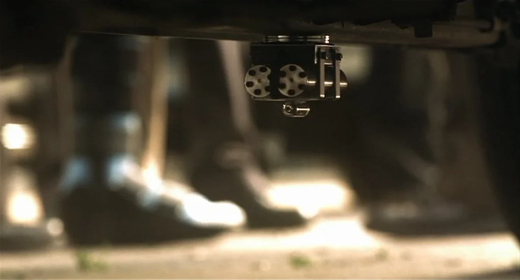 Traffic (2000) movie still: closeup — A metallic mechanical device is attached to the frame of a vehicle chassis. The blurred background shows feet…