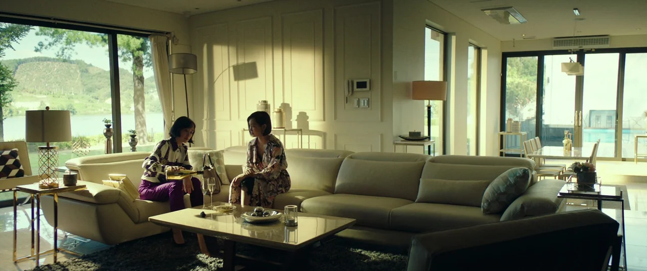 Beasts Clawing At Straws (2020) movie still: wide — A spacious, modern living room features large glass sliding doors and beige modular furniture.…