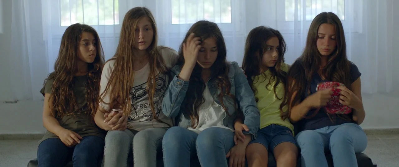 Mustang (2015) movie still: medium — Five teenage girls sit side-by-side on a dark couch against a windowed wall. They are positioned in a…