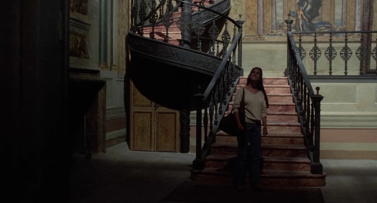 Etoile (1989) movie still: fullbody — An ornate interior staircase features a wrought iron railing and marble steps. A young woman stands on the…