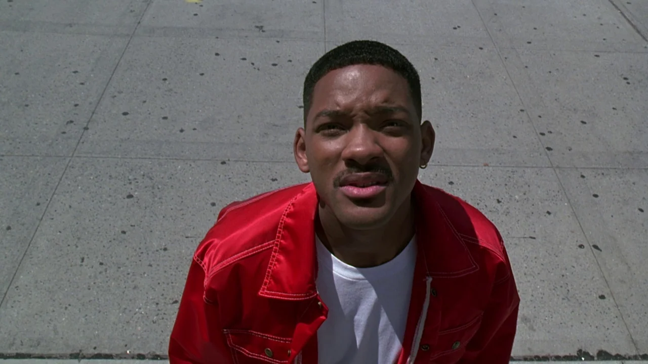 Men In Black (1997) movie still: medium — A grey concrete sidewalk fills the background of a high-angle shot. A man in a red jacket looks directly…