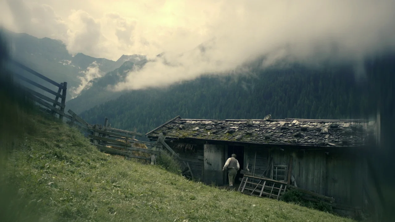 The Field Guide to Evil (2018) movie still: wide — A rustic wooden structure sits on a steep grassy slope near pine-covered mountains. A man walks…